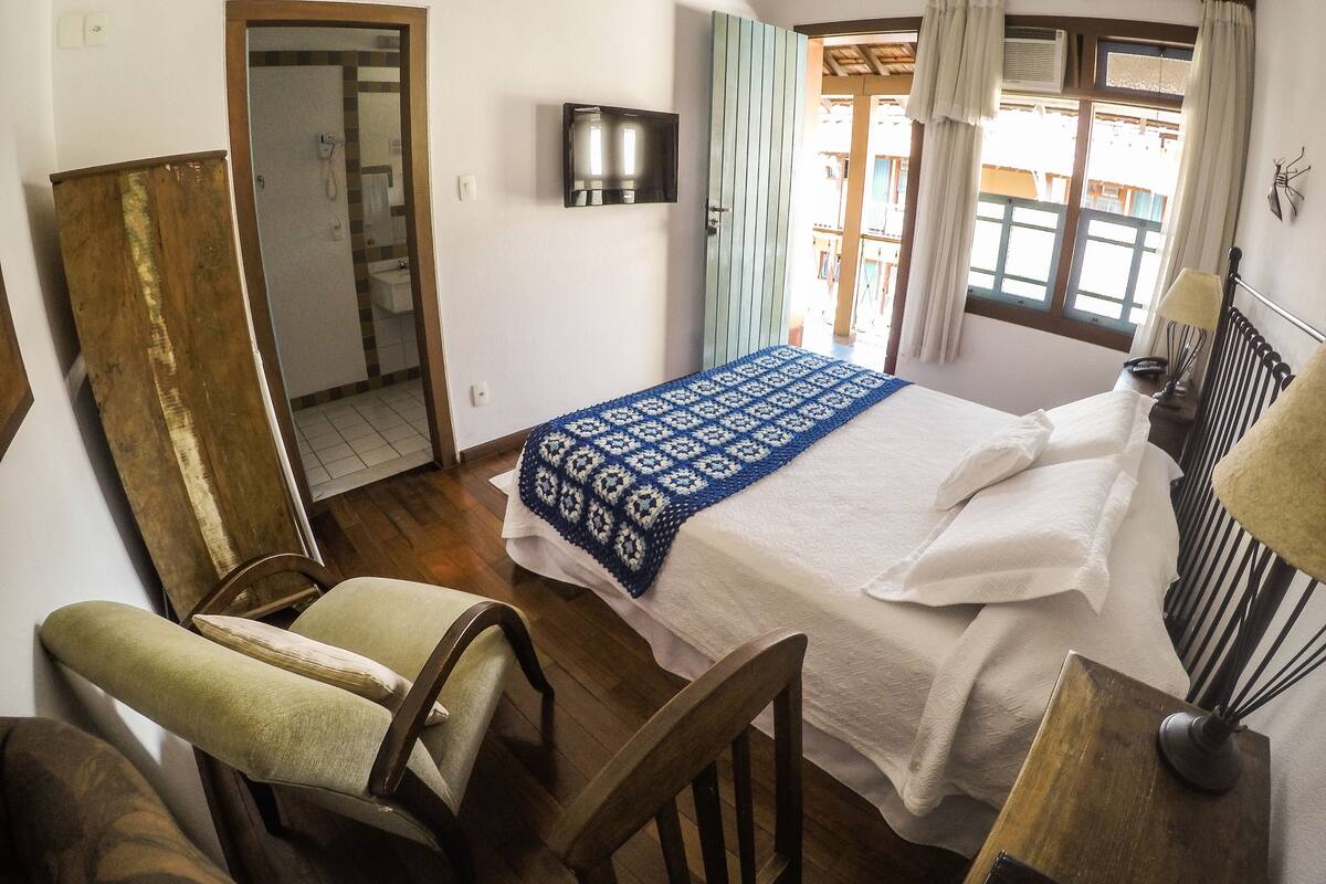 A bedroom with a bed, a chair, a wooden table, a TV, and a window with curtains.