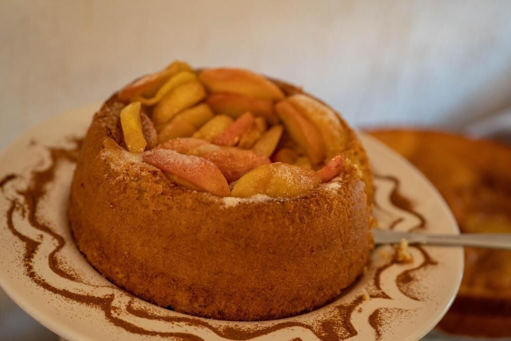 A round cake with sliced peaches on top, dusted with powdered sugar, on a plate with a decorative swirl pattern.