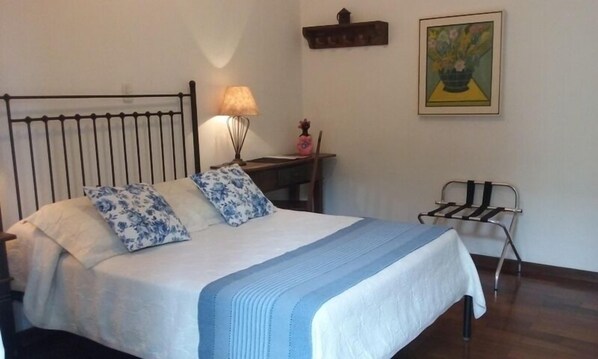 Una cama bien hecha con almohadillas florales, una lámpara de noche, una mesita con un florero y una silla.