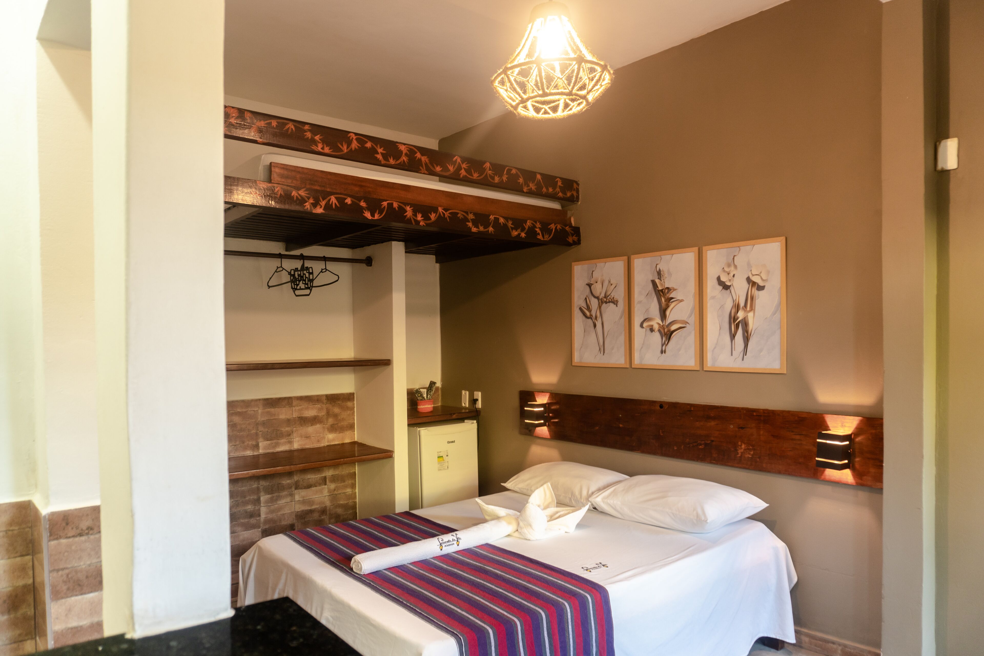 Une chambre d’hôtel avec un lit, une tête de lit décorée, une petite table avec une plante et un luminaire fixé au mur.