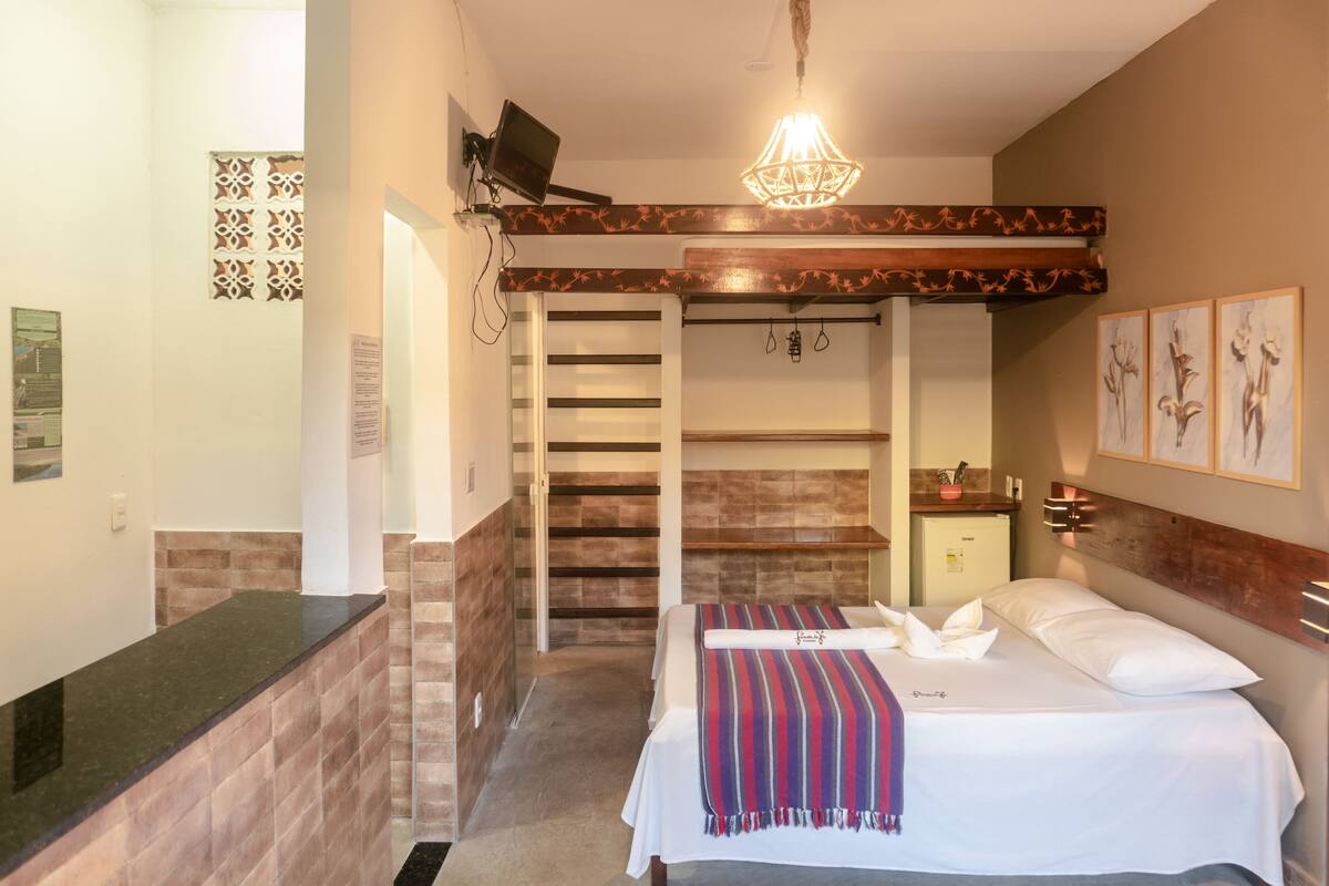 A hotel room with a bed, a small desk, a mirror, a wooden ceiling, and a staircase leading to another room.
