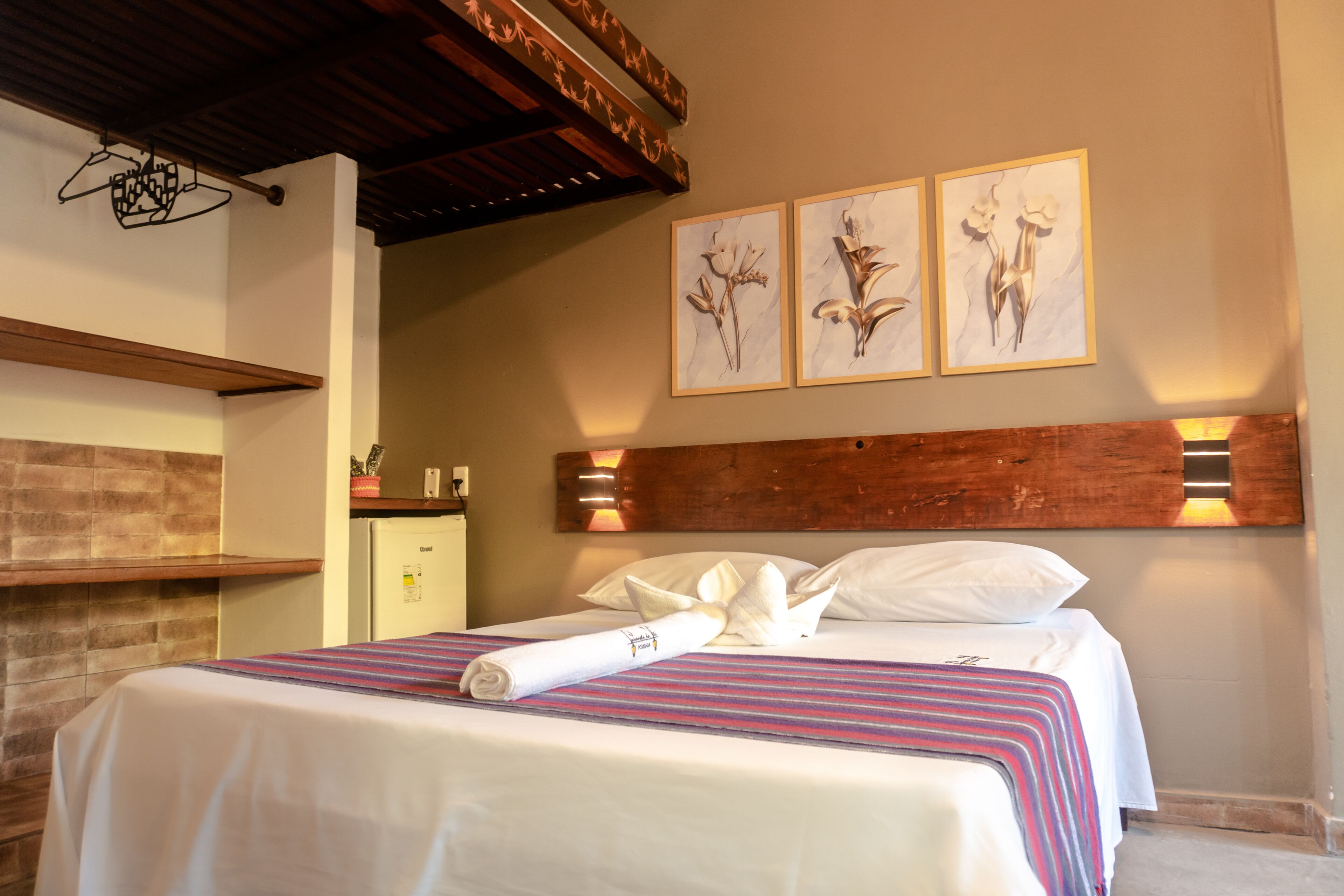 A neatly made bed with a striped bedspread, a rolled towel, and a wooden headboard. There are three framed floral artworks on the wall above the bed.