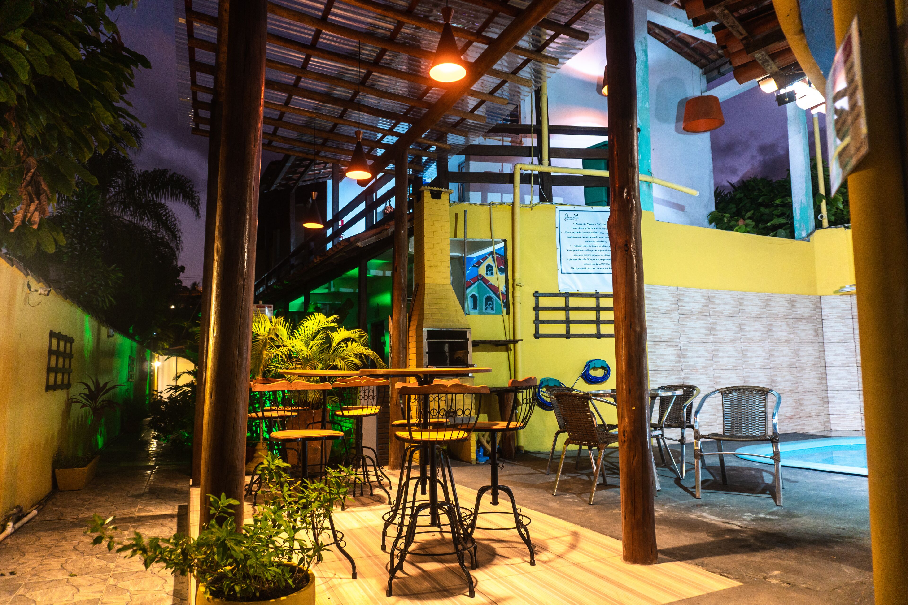 Un patio al aire libre con mesas y sillas, rodeado de plantas en macetas y una piscina.