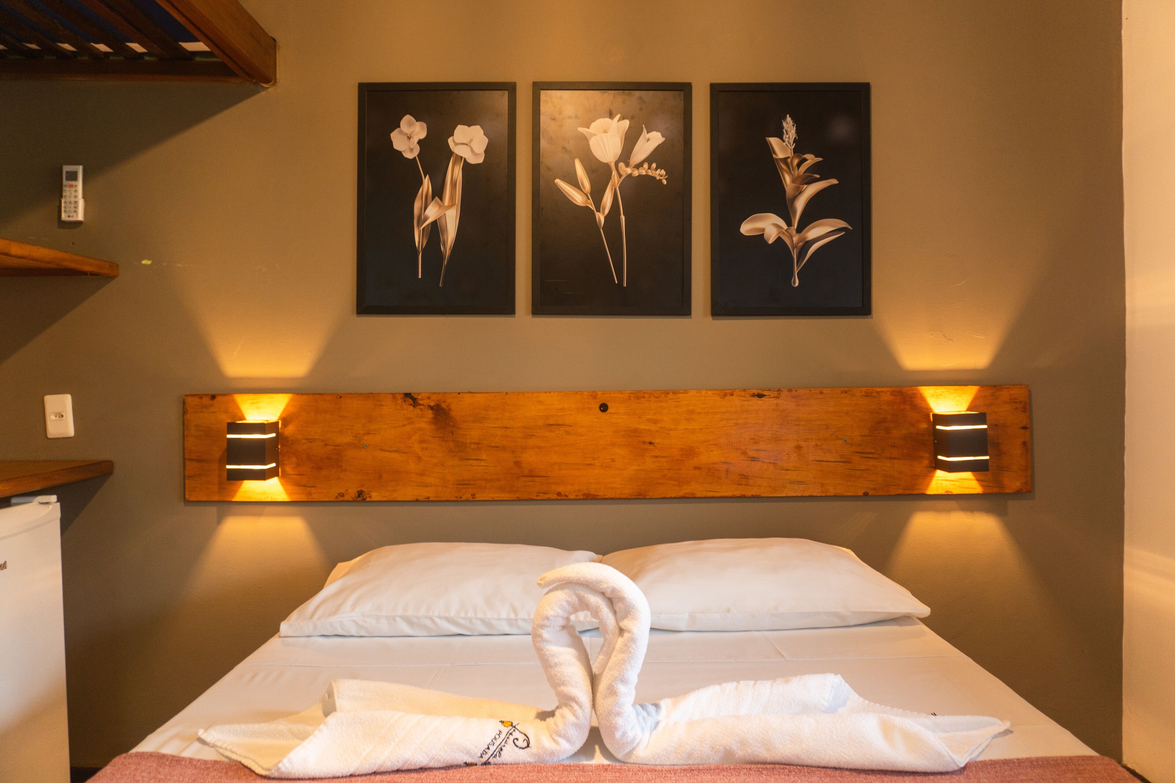 Quarto de hotel com cabeceira de madeira, cama com roupa de cama branca e três quadros florais emoldurados na parede.