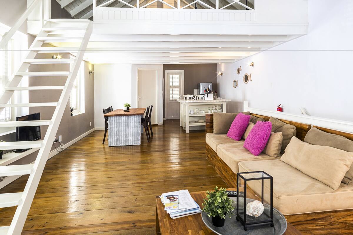 A spacious living area with a wooden floor, a beige sofa with pink cushions, a coffee table with magazines and a plant, and a staircase leading to an upper level.