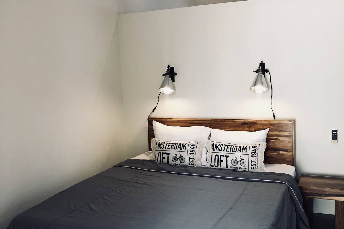 A modern bedroom with a wooden headboard, a grey bedspread, two wall-mounted reading lamps, and a nightstand with a lamp.