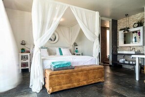 A bedroom with a four-poster bed, a bedside table, a nightstand, a bench, and a mirror above the sink. - Pietermaai Ocean Front (Willemstad)