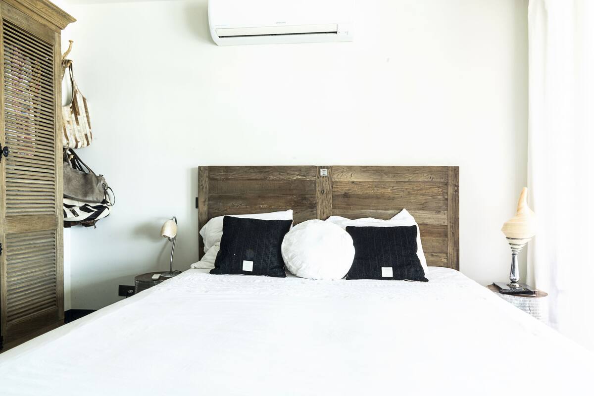 A neatly made bed with a wooden headboard, a bedside lamp, and a wall-mounted air conditioner.