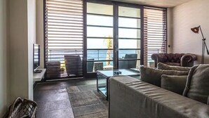 A living room with a sofa, a coffee table, and a lamp. There are large windows with blinds, a view of the ocean, and a balcony. - Pietermaai Ocean Front (Willemstad)