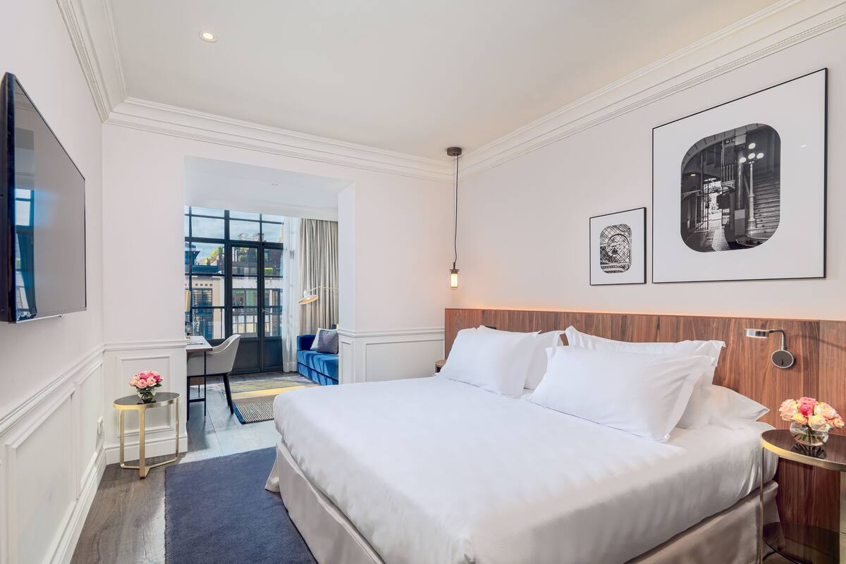 A hotel room with a large bed, a TV, a desk, and a view of the city through large windows.