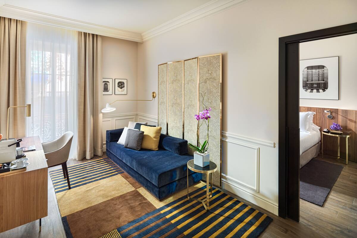 A hotel room with a blue sofa, a desk with a lamp, a TV, and a bed with a nightstand.
