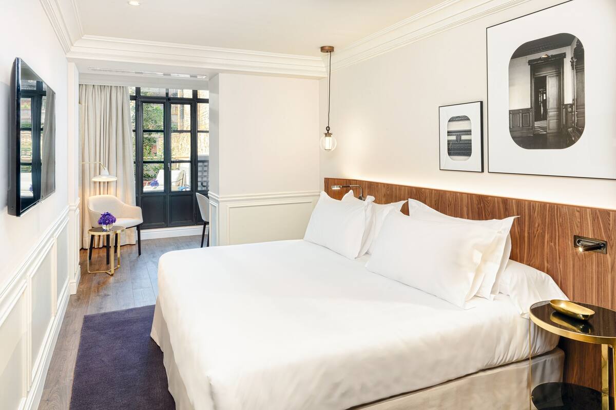 A hotel room with a large bed, a bedside table, a chair, a lamp, and a door leading to a balcony.
