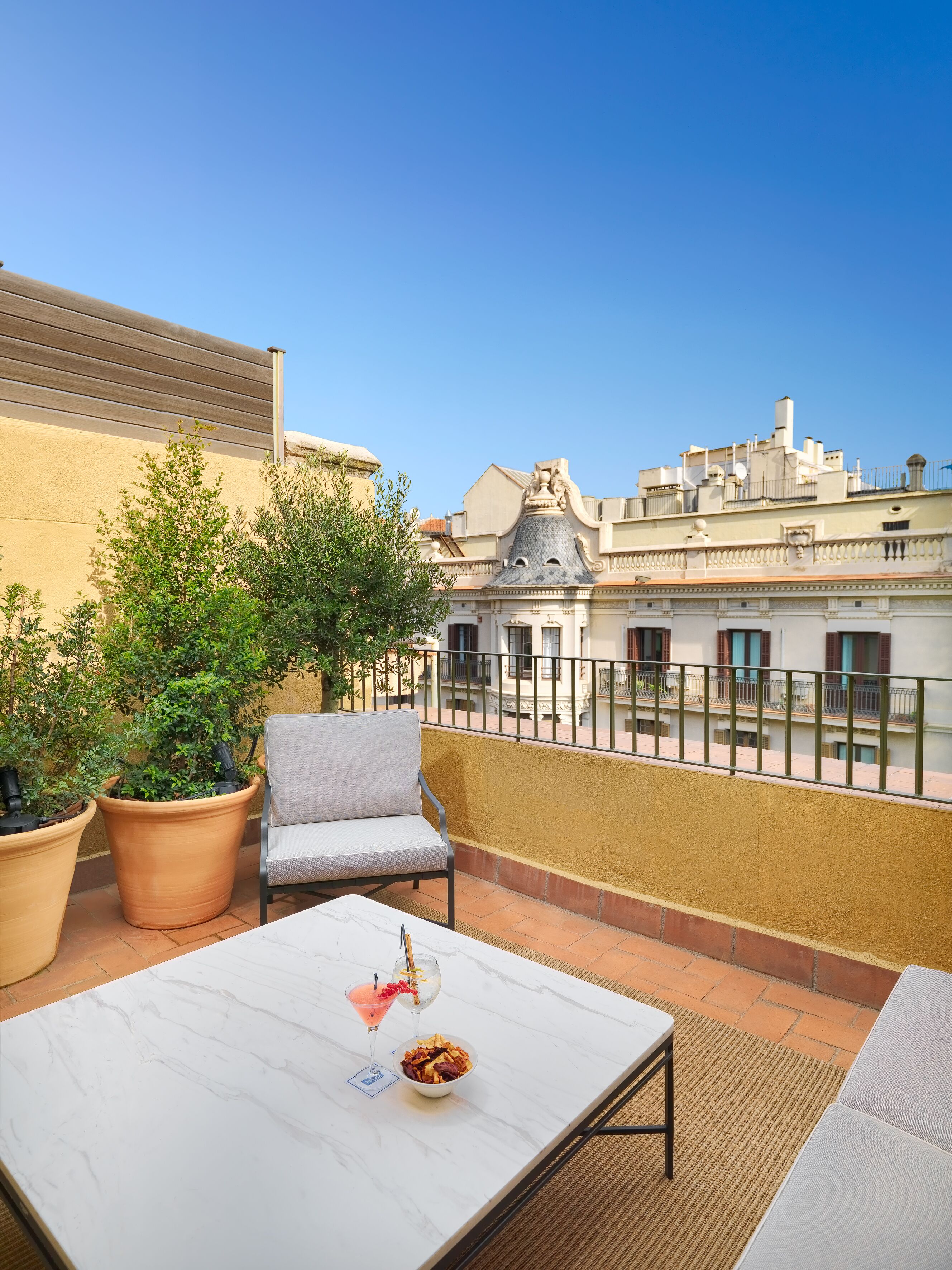 Una terraza con una mesa de mármol blanco, una silla gris, plantas en macetas y vistas de edificios al fondo.