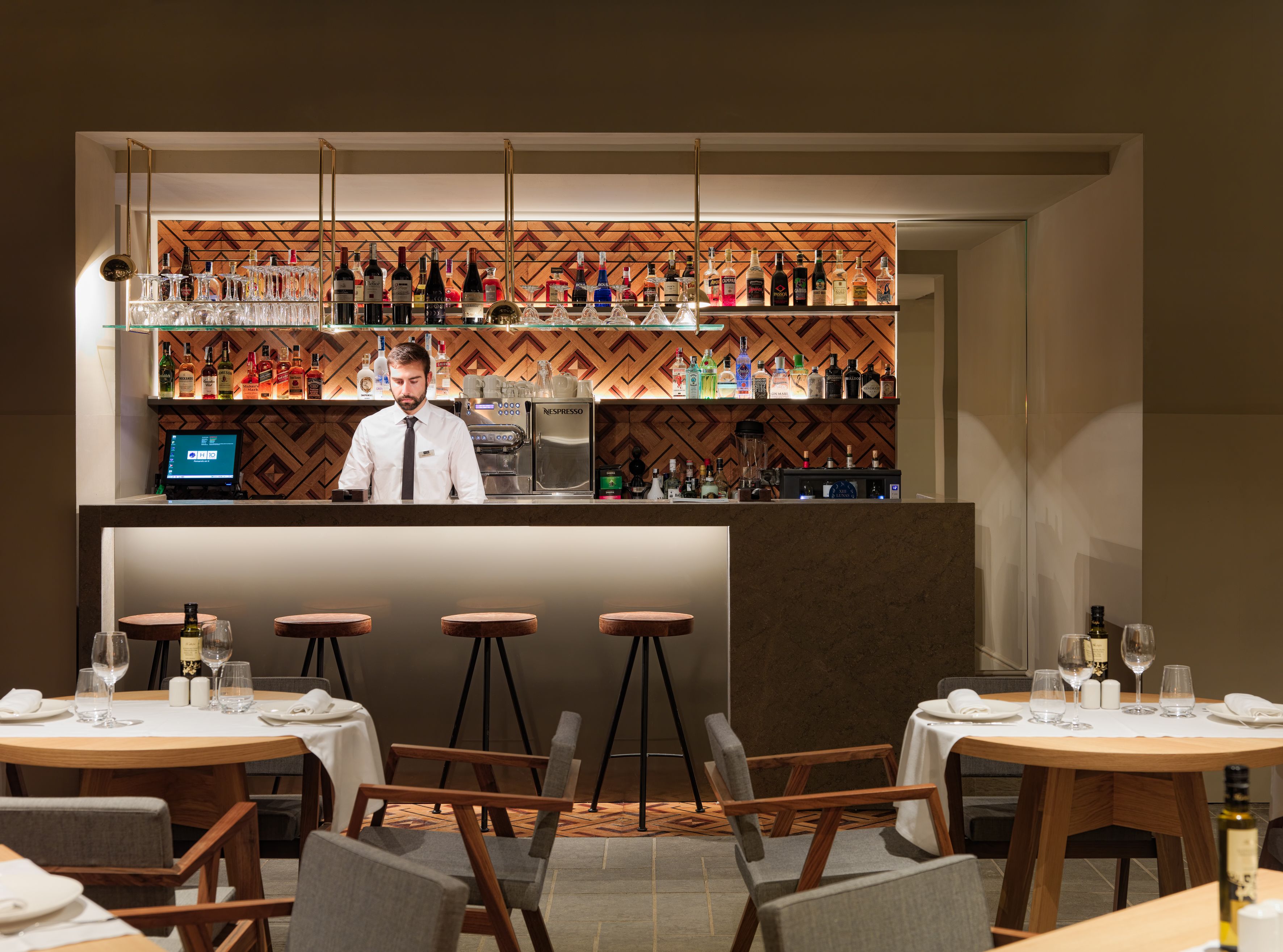 A bar area with a bartender, a coffee machine, and a variety of bottles and glasses on shelves.