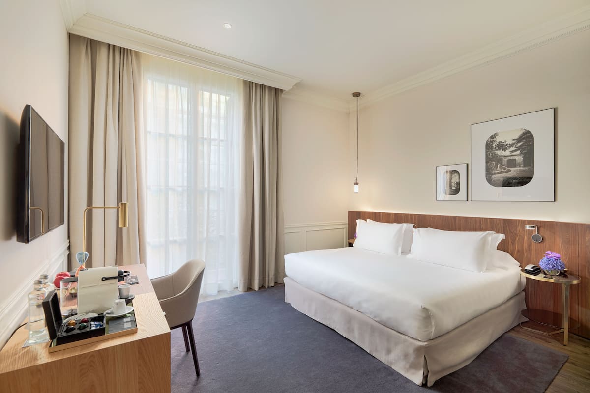 A hotel room with a large bed, a desk with a chair, a TV, and a bedside table with a lamp.