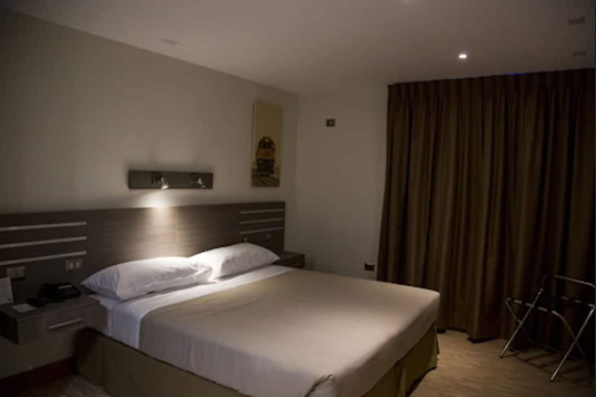 Ein Hotelzimmer mit einem großen Bett, einem Nachttisch, einer Lampe, einem Vorhang und einem kleinen Tisch.