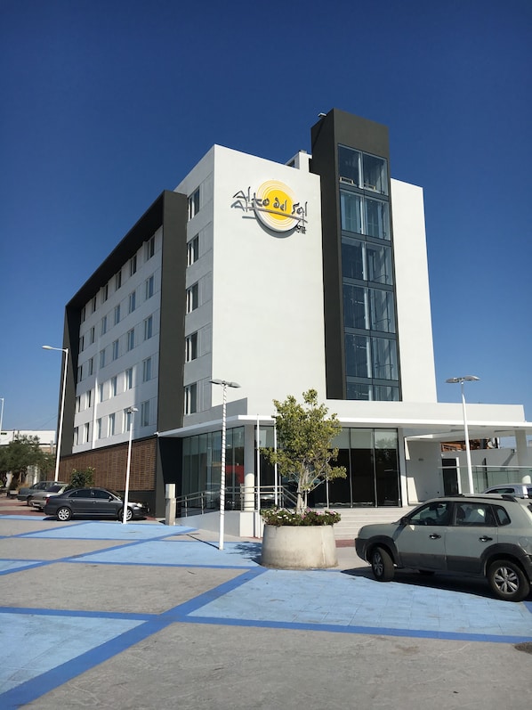 A modern hotel building with a prominent logo on the facade, large windows, and a parking area with several cars. - Hotel Alto del Sol Plaza Mejillones (Mejillones)