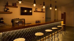 A bar area with a long counter, multiple stools, hanging pendant lights, and shelves with bottles.