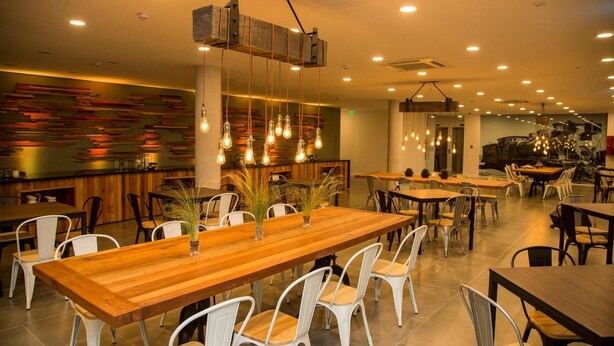 A modern restaurant interior with wooden tables and white chairs, hanging light fixtures, and a colorful abstract wall design.