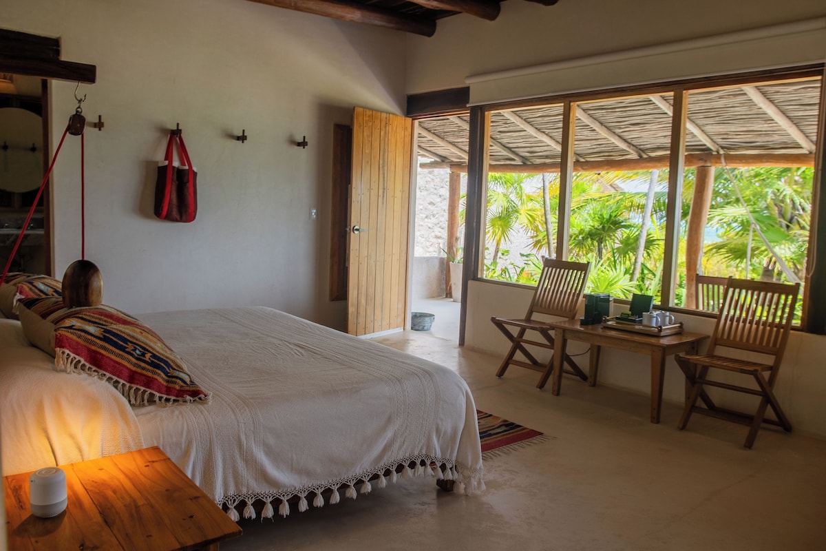 A bedroom with a bed, a nightstand, a table with chairs, and a view of palm trees through large windows.