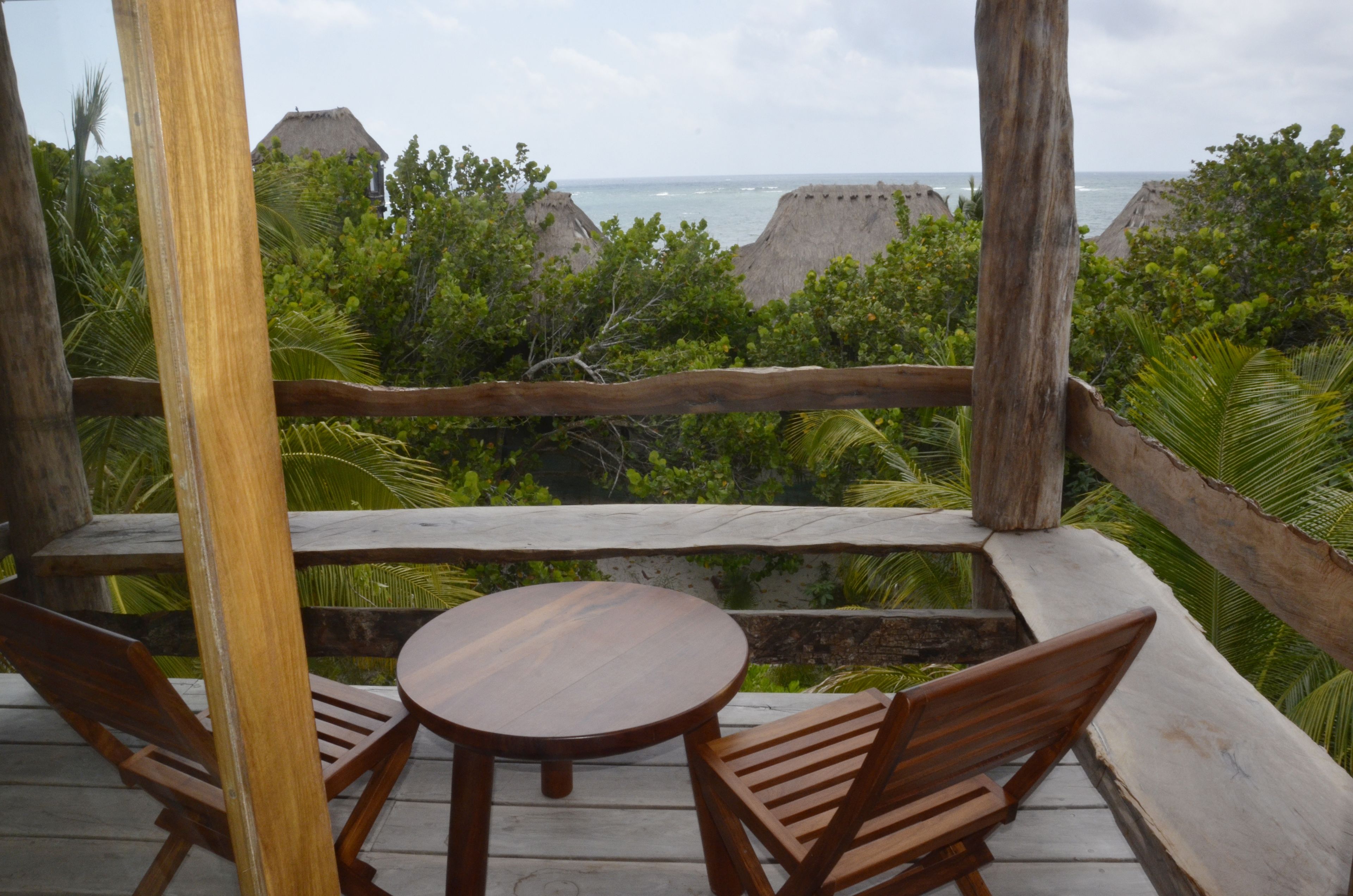 A wooden deck with a table and chairs, overlooking a lush landscape with trees and a view of the ocean.