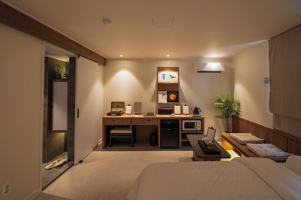 A hotel room with a bed, a desk with a computer, a microwave, a small fridge, and a bedside table with a lamp and a clock.
