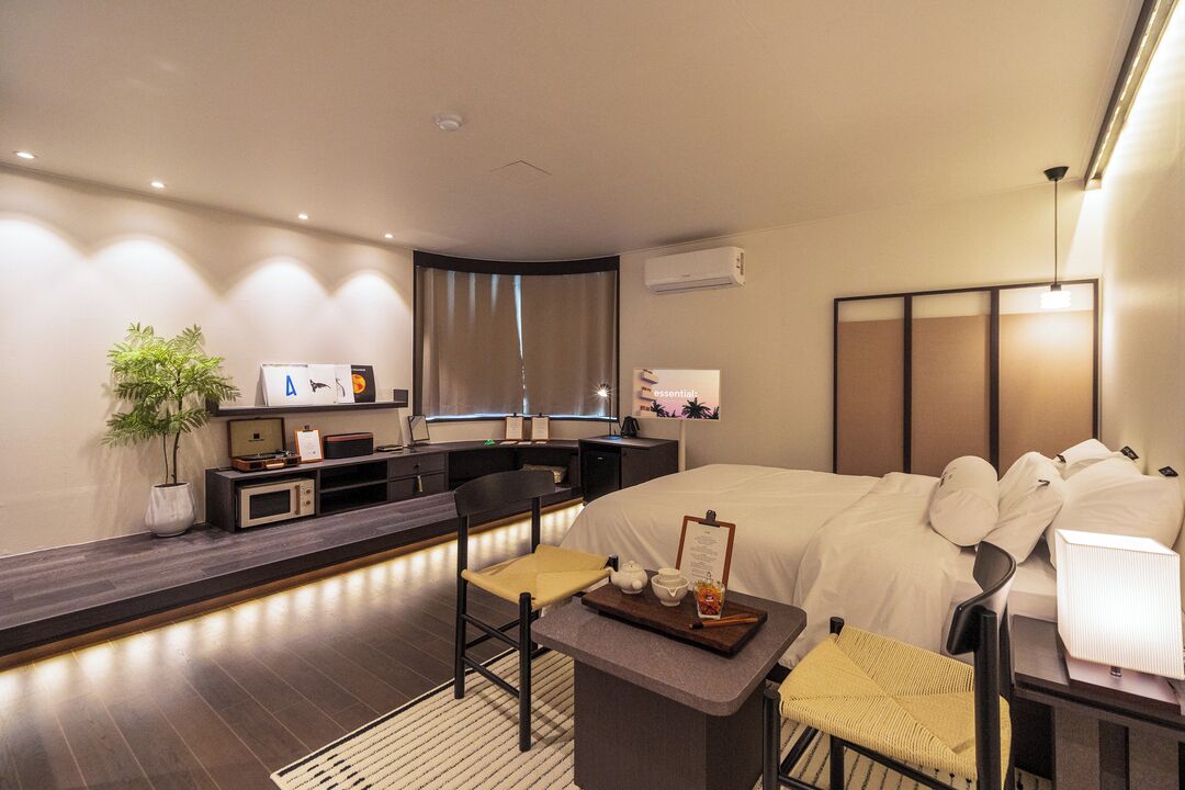A modern hotel room with a bed, bedside table, chair, desk, and a small plant.