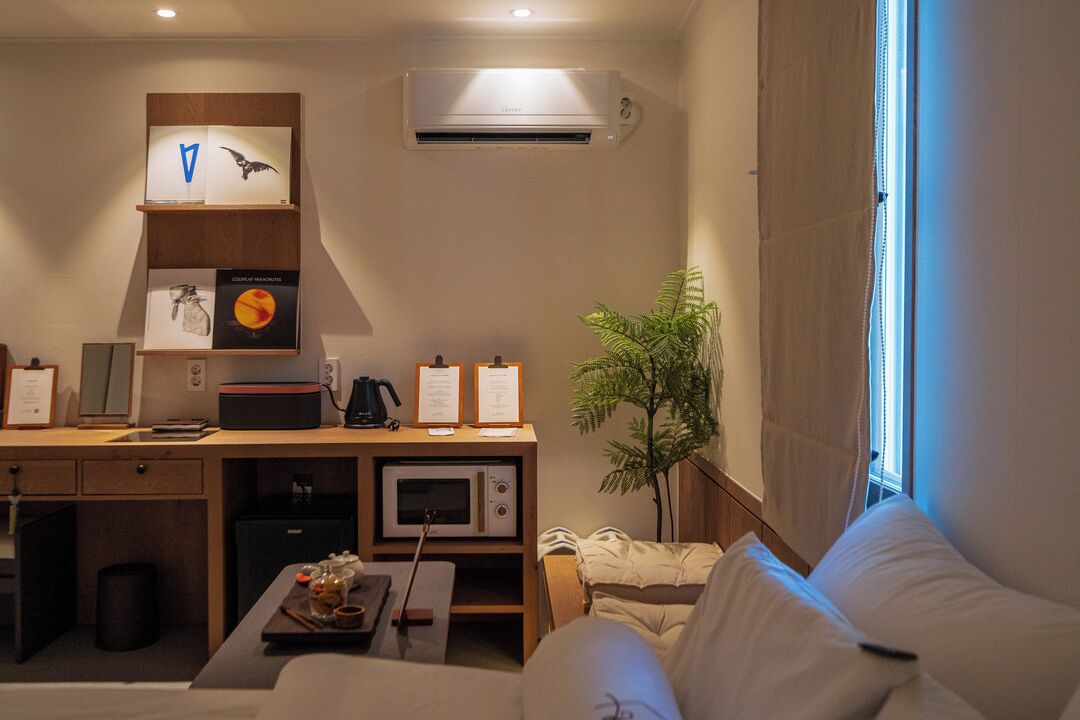 A hotel room with a bed, a desk with a computer, a microwave, a small fridge, a plant, and a window with curtains.