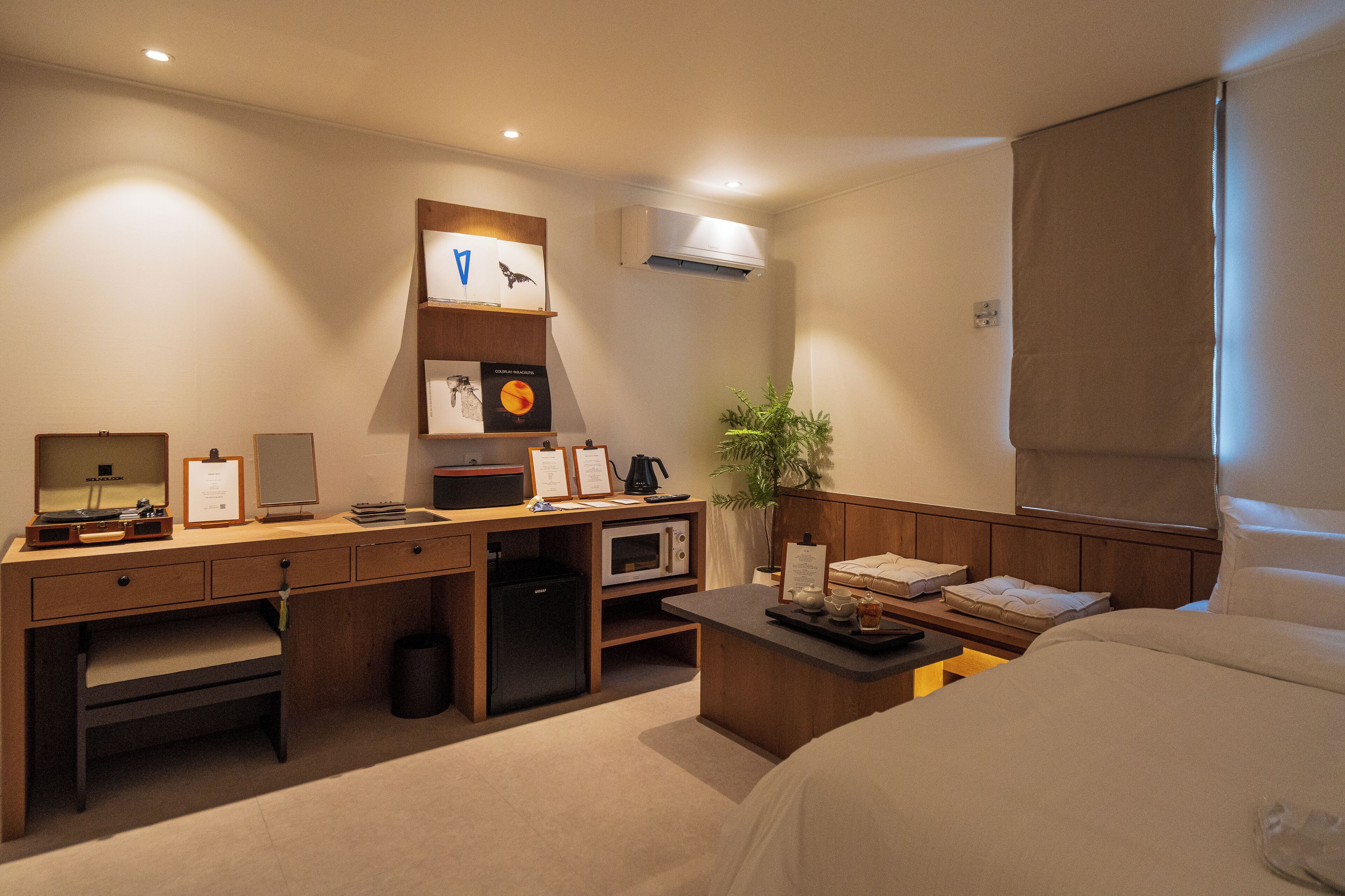 Habitación de hotel con cama, escritorio con computadora, microondas, un pequeño basurero y una planta en maceta.