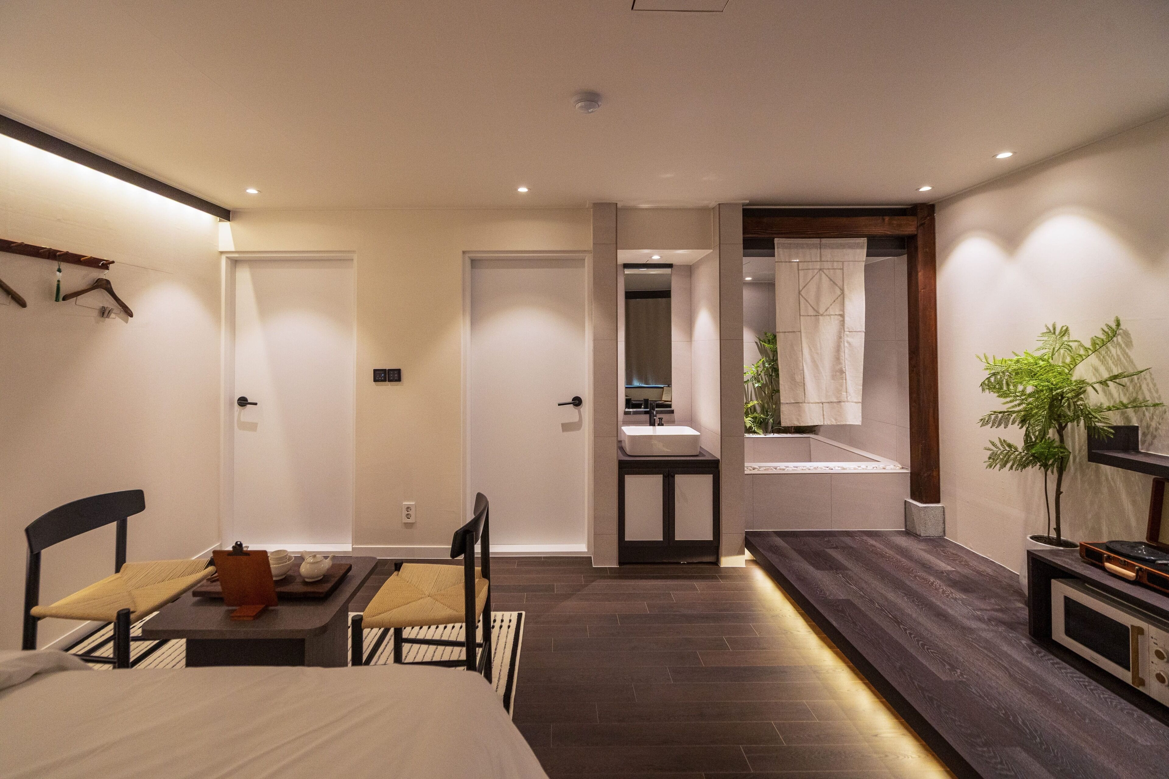 Una camera d'albergo moderna con un letto, comodino, sedia e un piccolo angolo ristoro. È presente un bagno con lavandino e specchio.