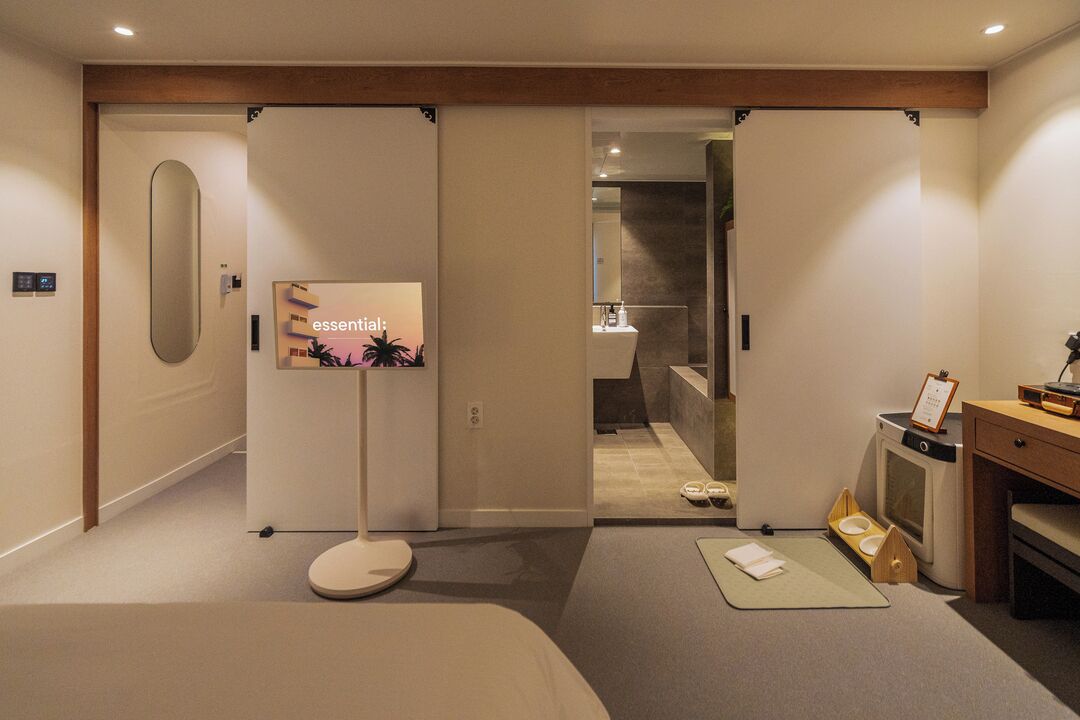 A modern hotel room with a sliding door leading to a bathroom, a flat-screen TV, a desk with a chair, and a small seating area.