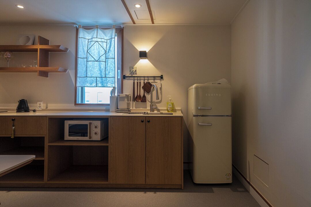 A compact kitchen with a microwave, a refrigerator, wooden cabinets, a window with a view, and a wall-mounted light fixture.