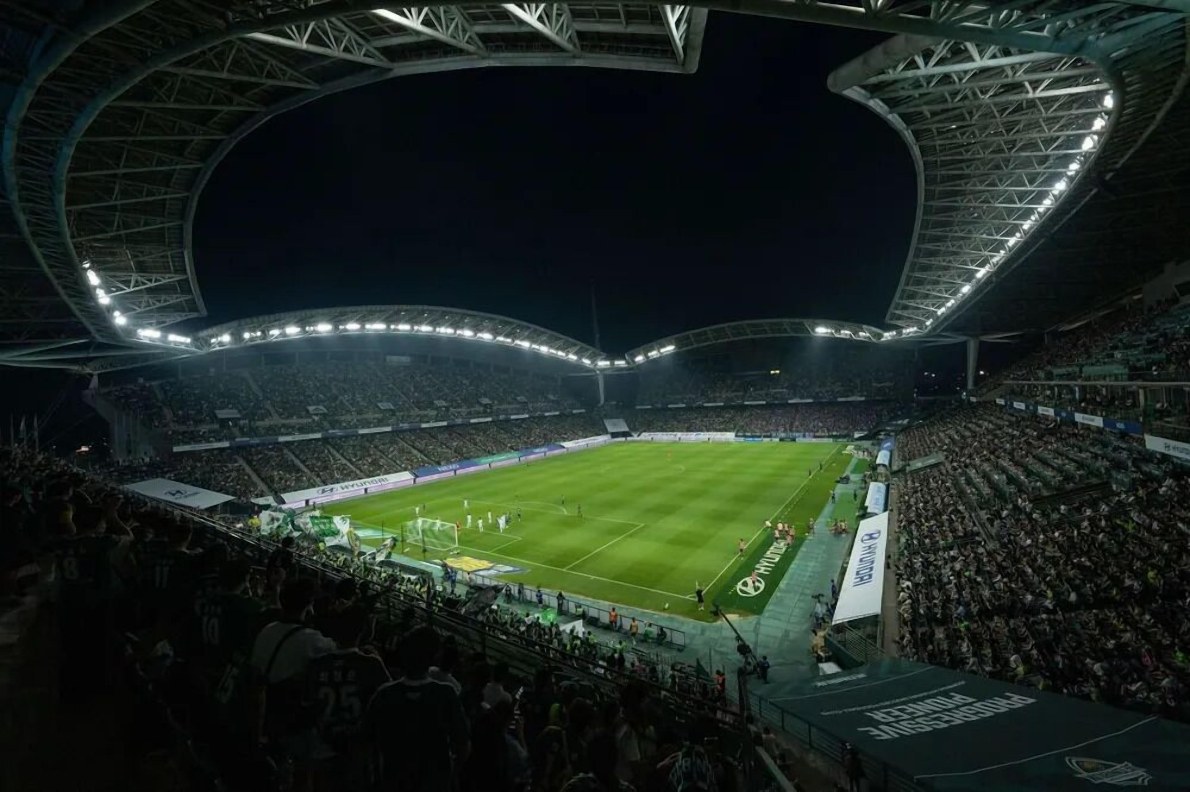 A large stadium with a curved roof structure, multiple tiers of seating, and a well-lit field.