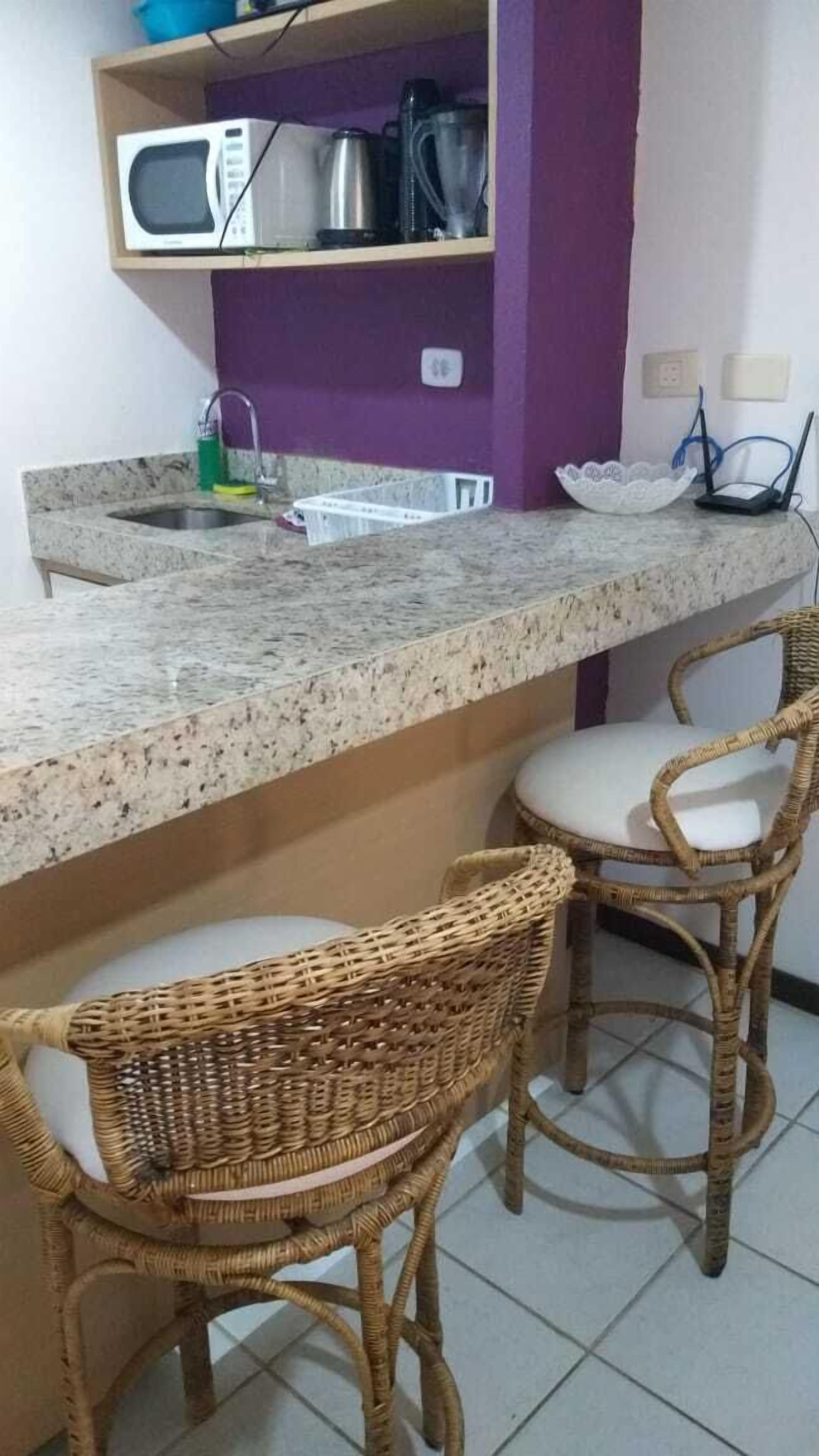 Cozinha com bancada de granito, micro-ondas, chaleira e liquidificador em uma prateleira roxa. Há duas cadeiras de vime com almofadas brancas.