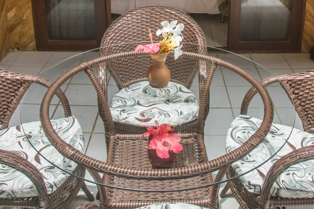 A room with a bed, a wicker chair, and a table with a vase of flowers.