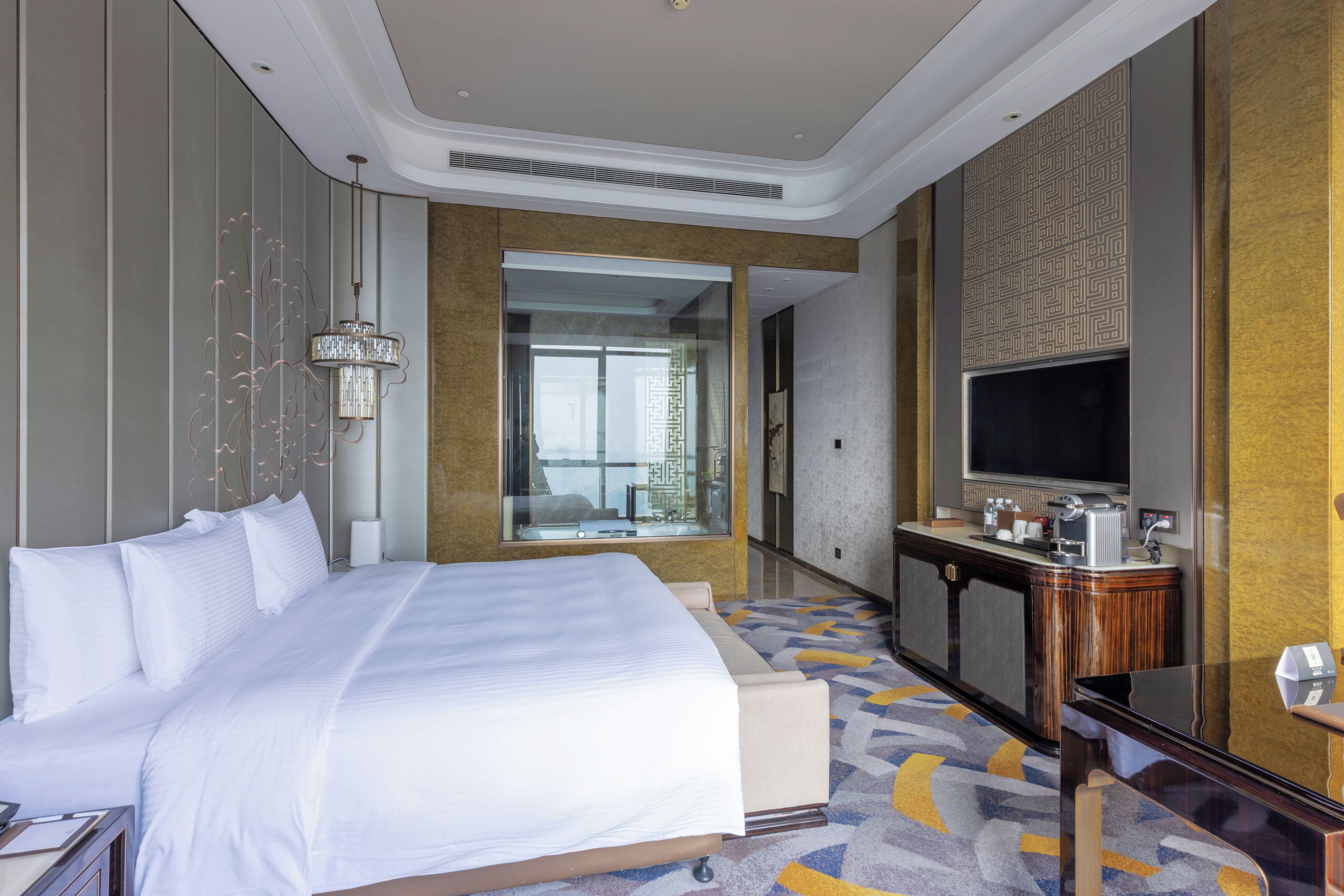 A hotel room with a large bed, a desk with a flat-screen TV, a minibar, and a view of the city through the window.