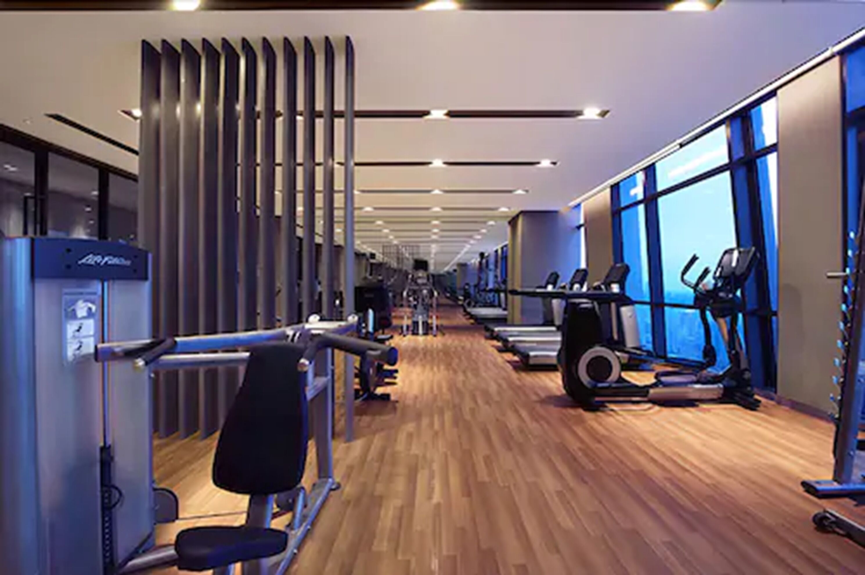 A modern gym with cardio machines, weight stations, and large windows providing natural light.