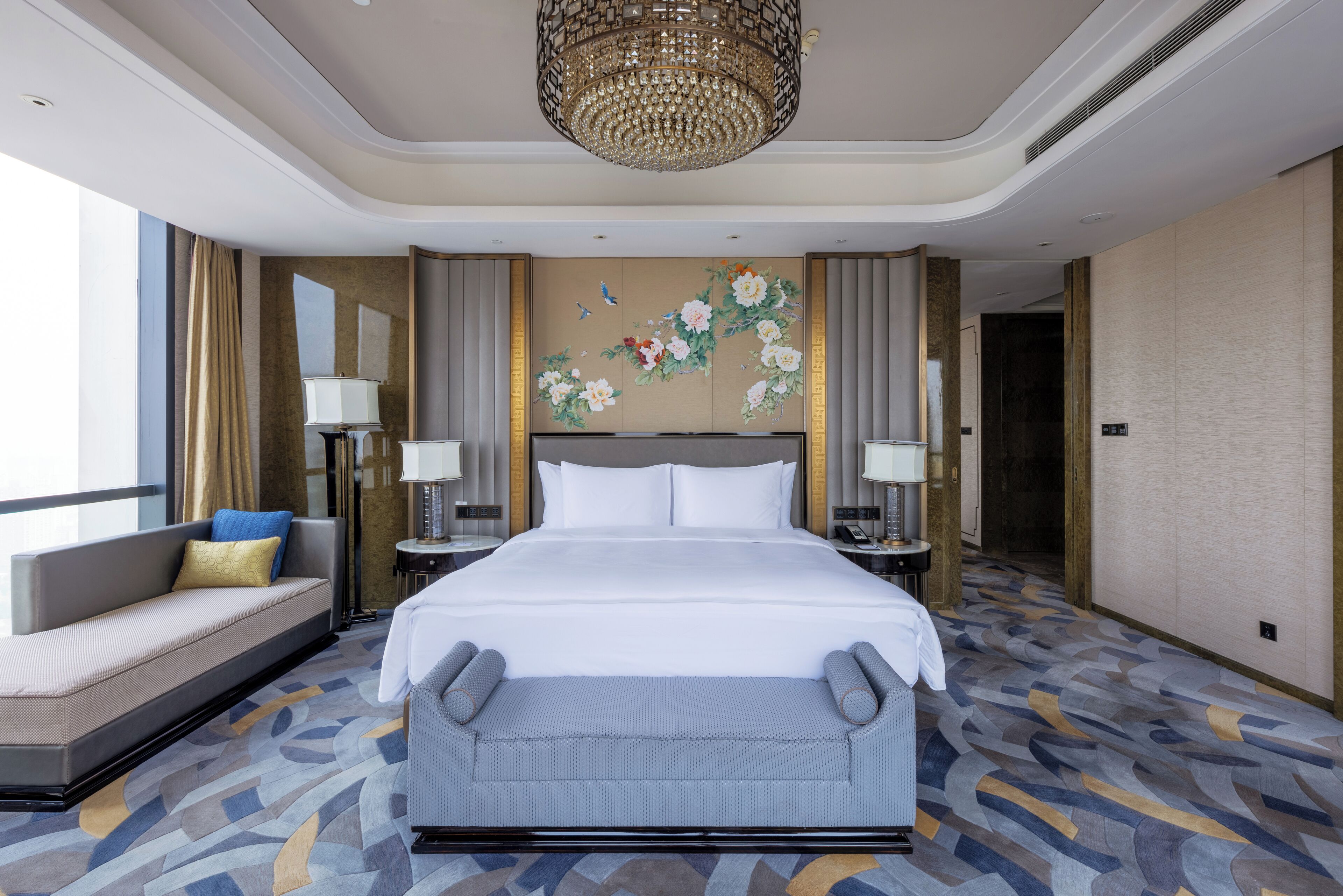 A hotel room with a large bed, a sofa, two nightstands, a chandelier, and a floral wall mural.