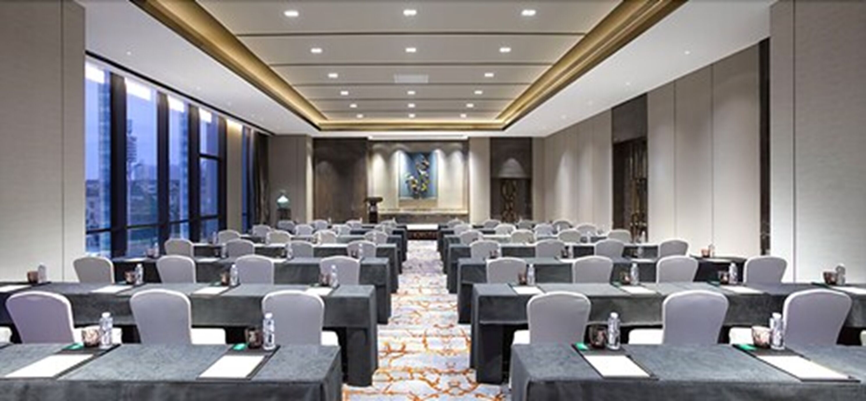 A conference room with rows of tables and chairs, large windows, and a carpeted floor.