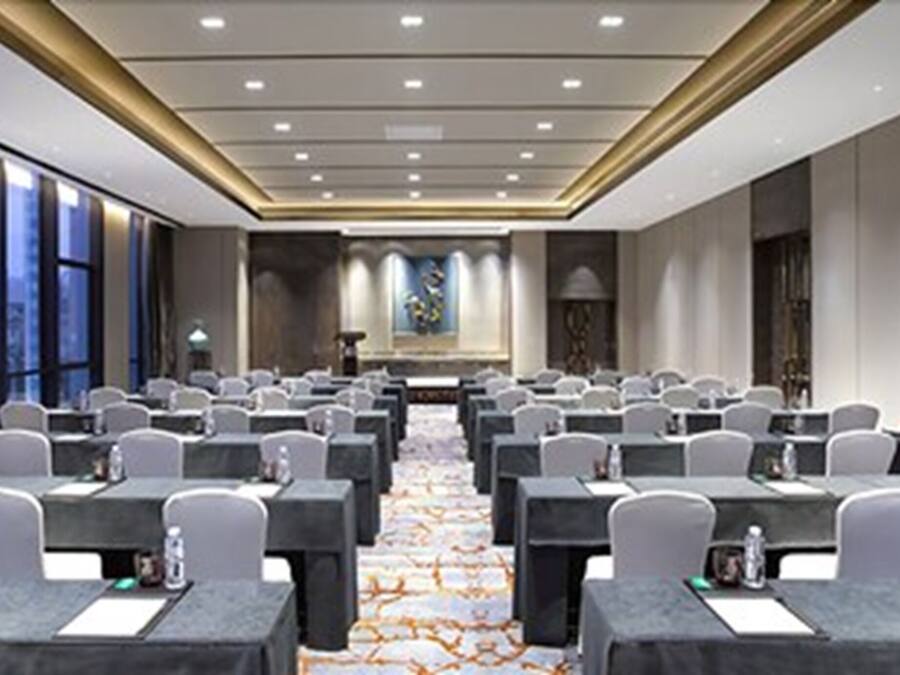A conference room with rows of tables and chairs, large windows, and a carpeted floor.