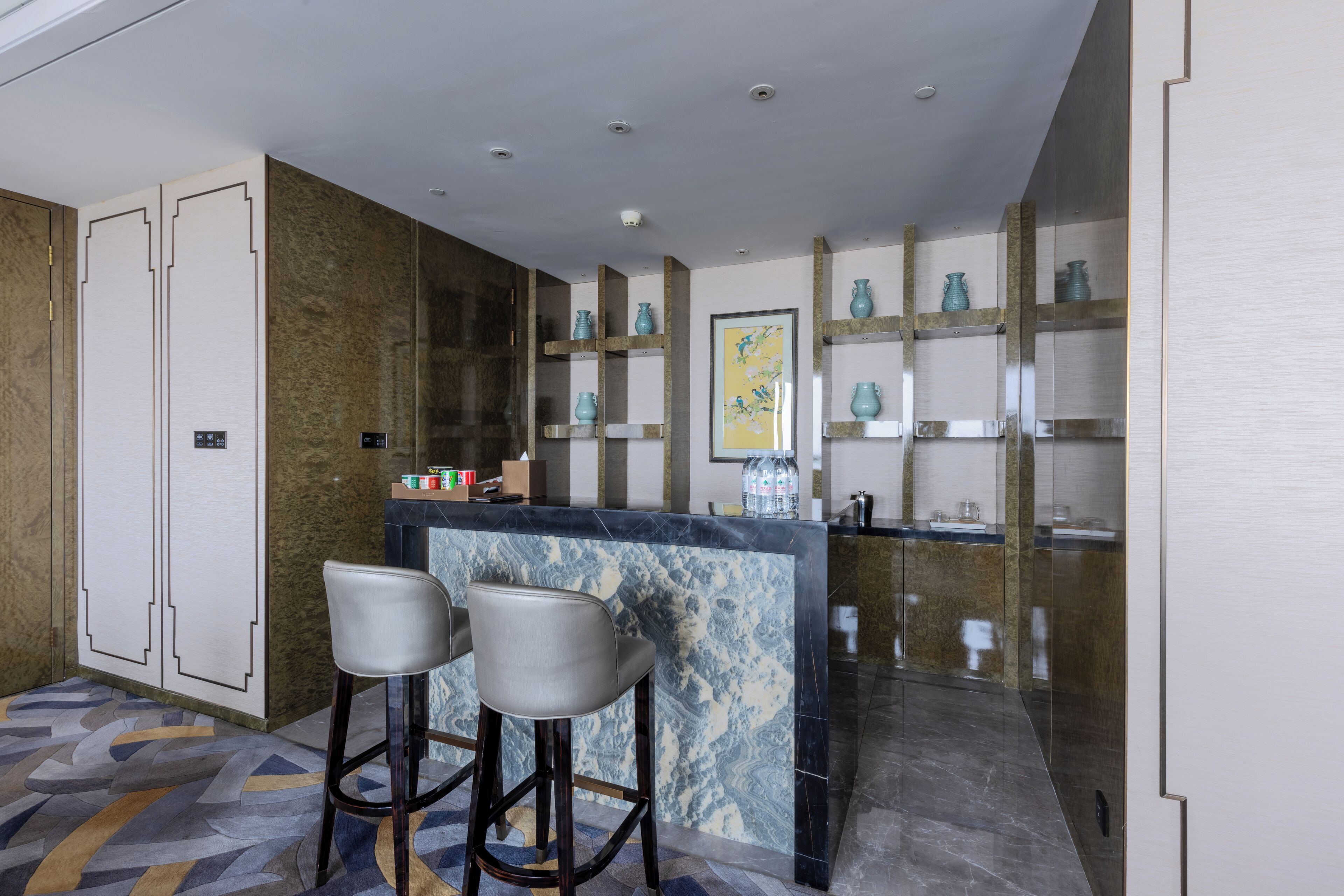 A modern kitchen with marble countertops, two bar stools, and decorative vases on shelves.