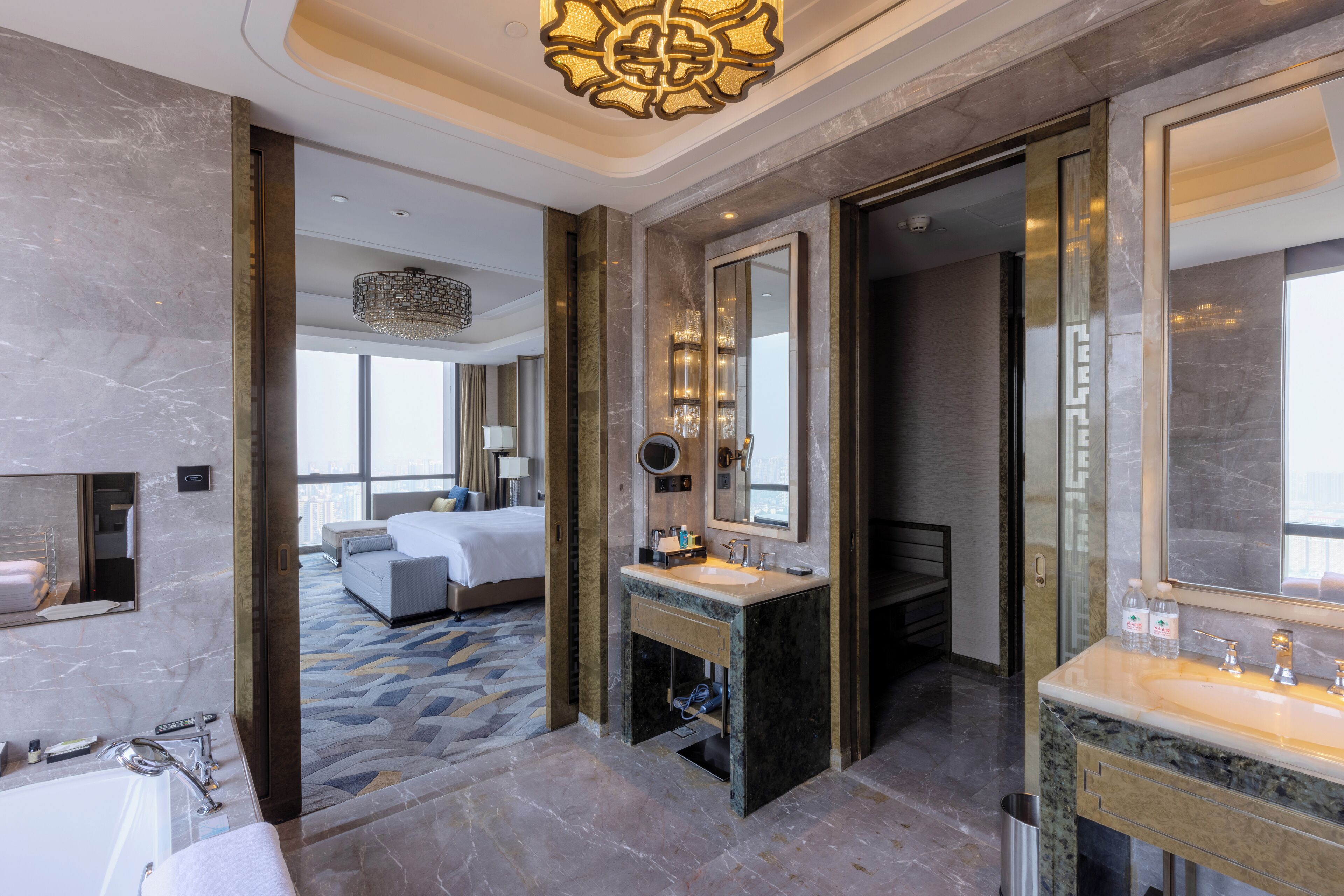 A modern hotel bathroom with a large bathtub, a sink, and a mirror. There is a bed visible in the background.