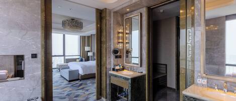 A modern hotel bathroom with a large bathtub, a sink, and a mirror. There is a bed visible in the background.