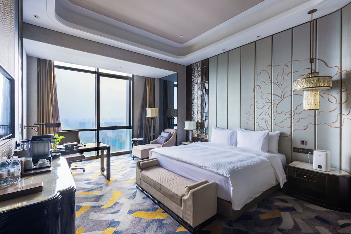 A hotel room with a large bed, a desk with a chair, a sofa, a nightstand with a lamp, and a view of the city.