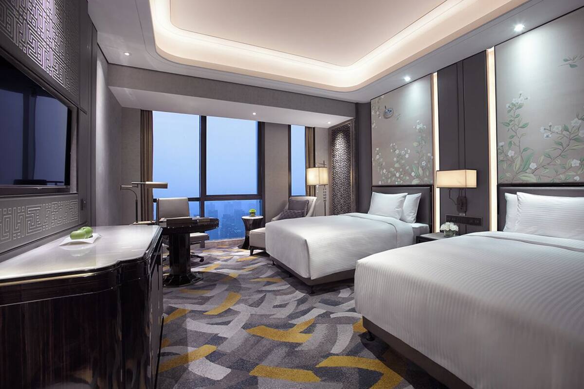 A hotel room with two beds, a desk, a chair, and a large window offering a city view.