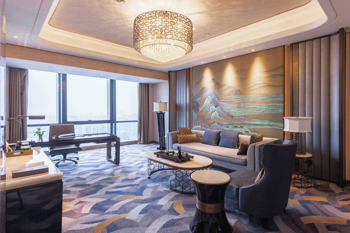 A modern hotel room with a large window, a sofa, a desk, and a coffee table.