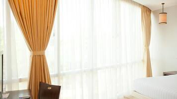 A hotel room with a bed, a desk with a chair, and a large window with sheer curtains.