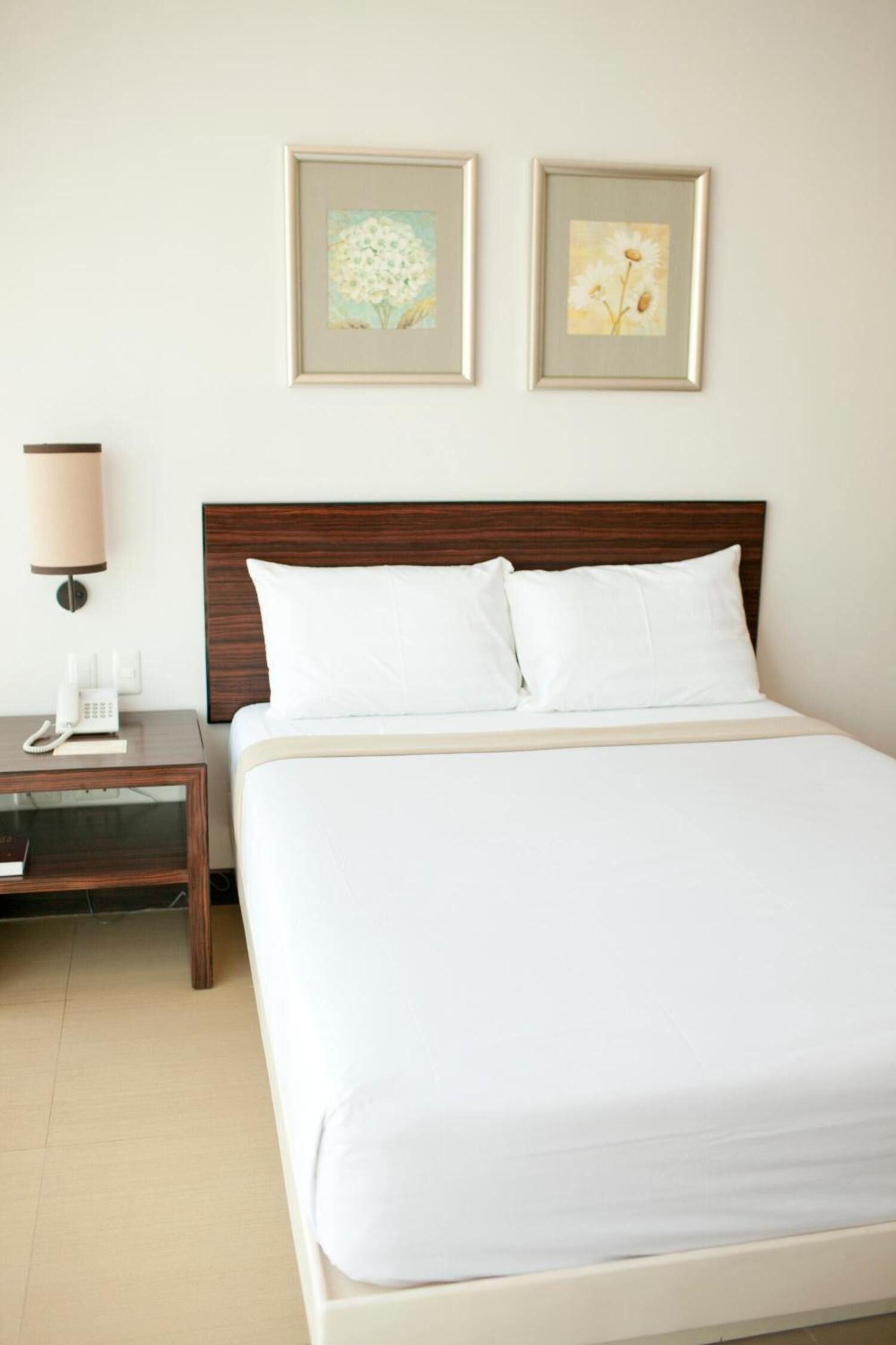 A hotel room with a bed, a nightstand with a lamp, and two framed floral paintings on the wall.