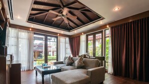 A spacious living room with a ceiling fan, large windows with curtains, a sectional sofa, and a coffee table. - Jantra Residence by Chinocollection (Kathu)