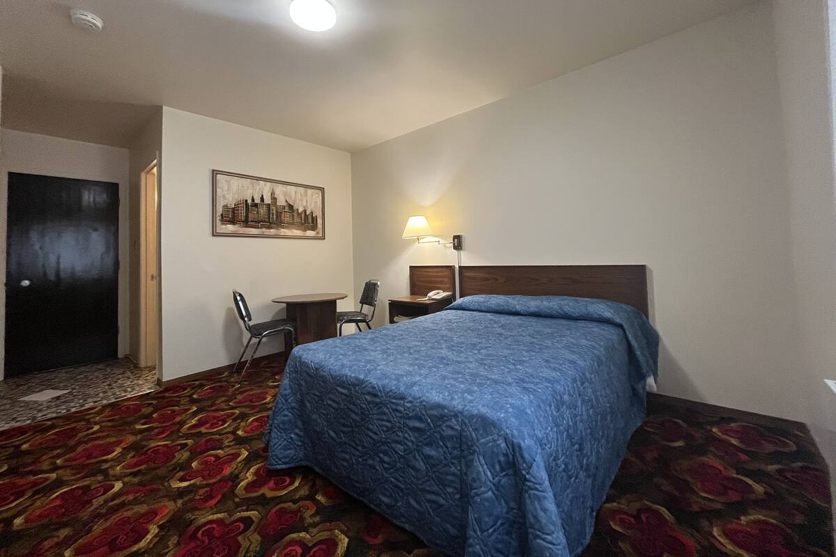Una habitación de hotel con cama, escritorio, silla, lámpara y un cuadro con una vista urbana.